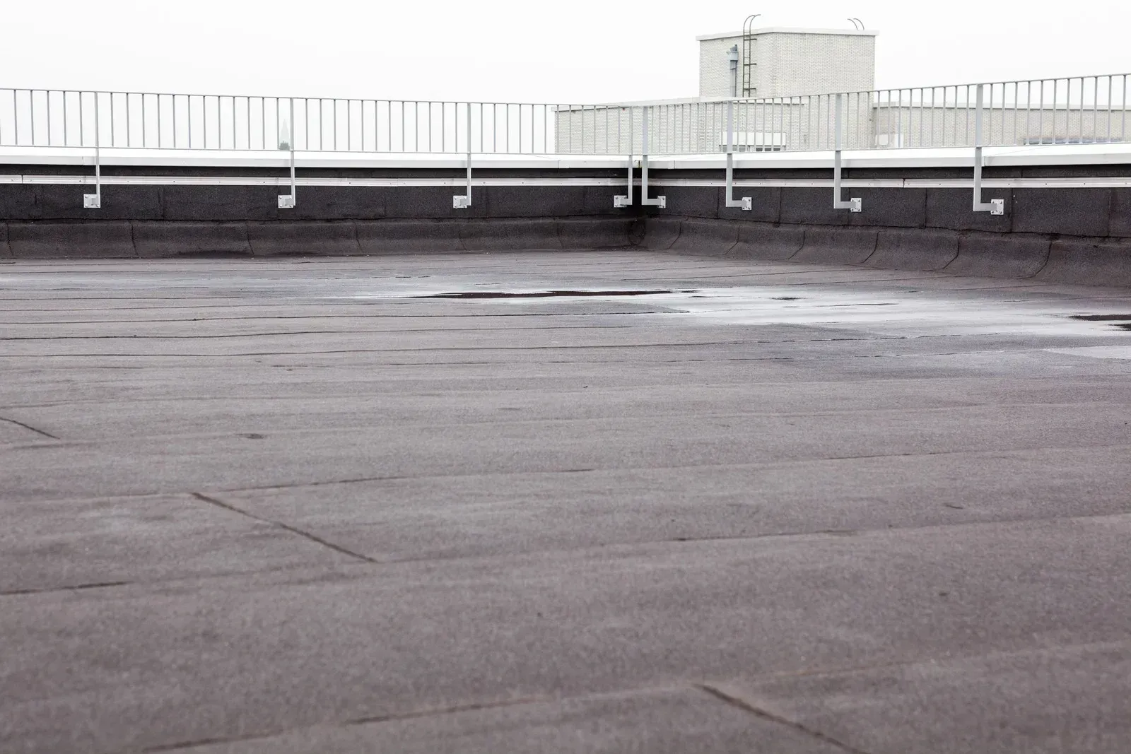 Flat, black, tar roof with metal railings and a distant building in a cloudy setting.