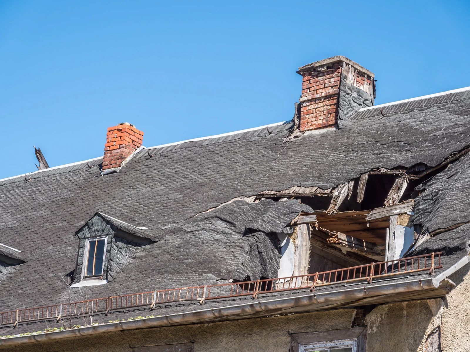 Damaged roof with missing sections, two brick chimneys, and a dormer window against a blue sky.