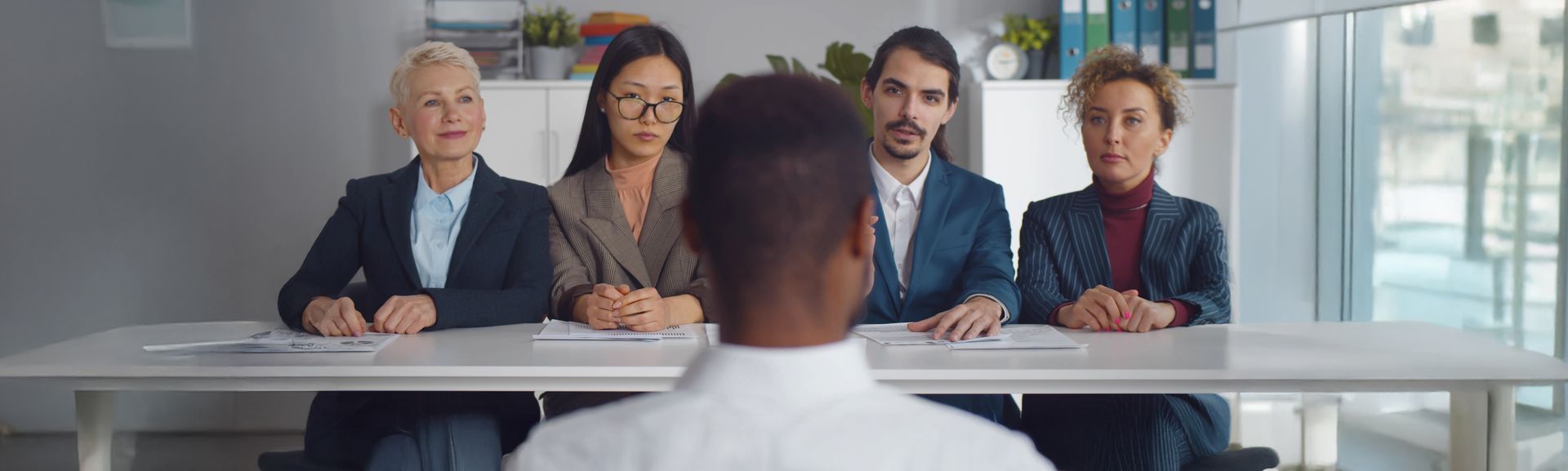 Image contenant un homme assis au premier plan devant une table d'entretien d'embauche devant une équipe de 4 recruteurs.