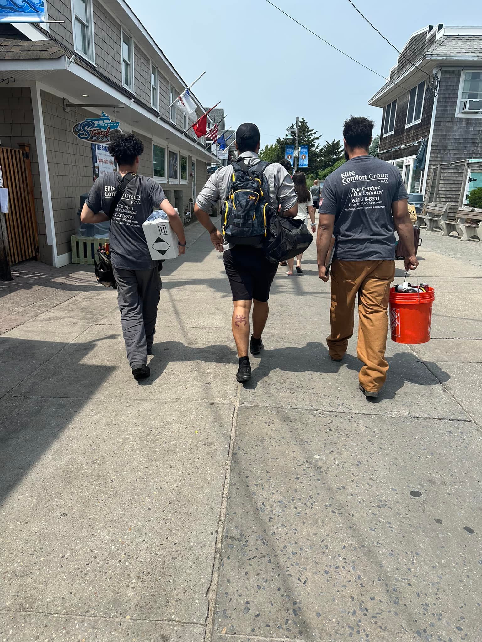 Three people walk away from the camera down a narrow, sunlit street between buildings, carrying bags and a red bucket.