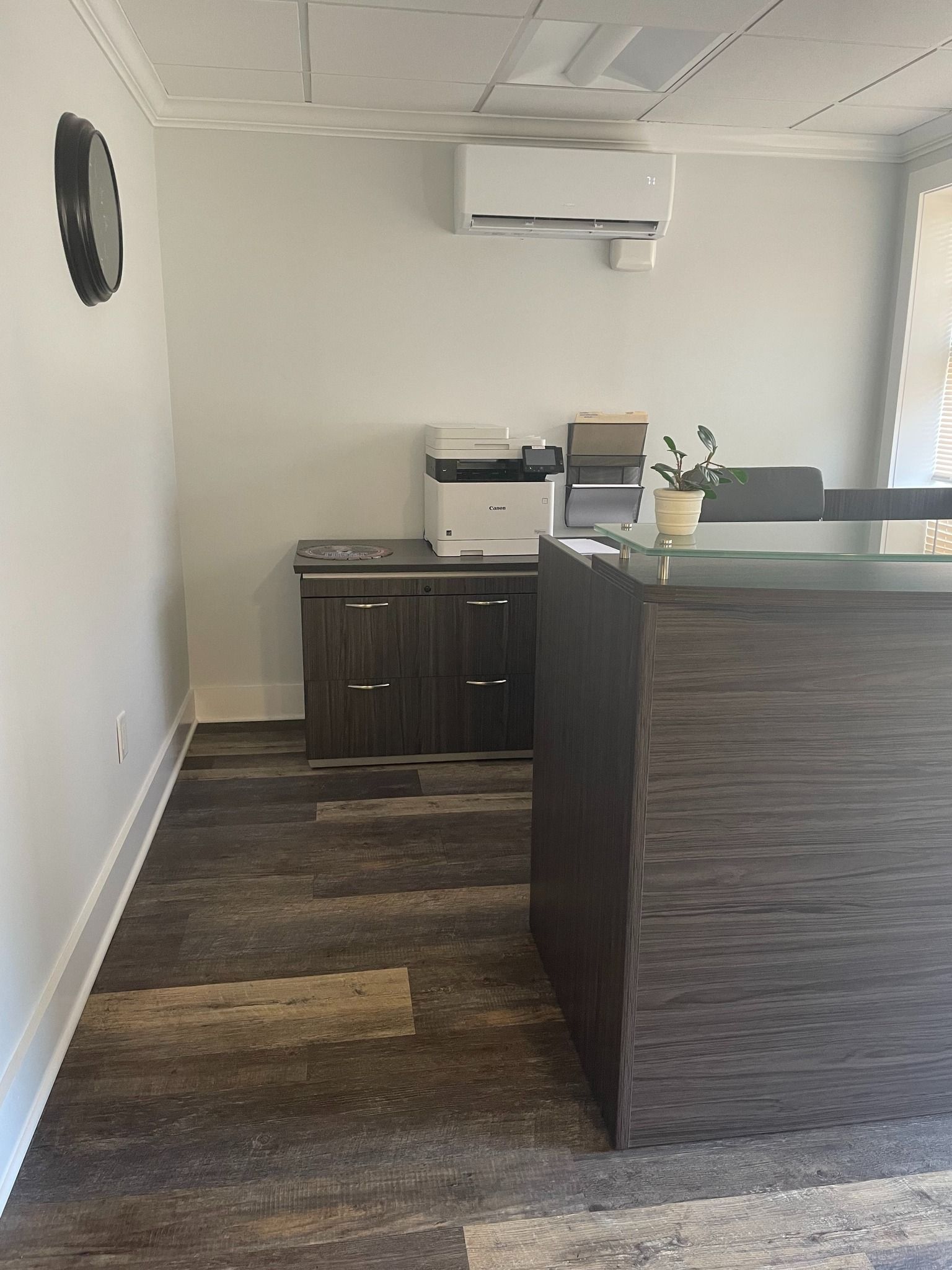 A modern office reception area with a dark wood desk, a printer on a matching cabinet, white walls, and wood-look flooring.