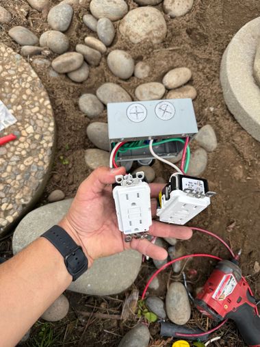A person uses a flashlight on an electrical panel at night, street and houses in background.