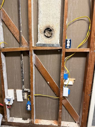 A person points at electrical wiring as another looks at a blueprint in a room with unfinished walls.