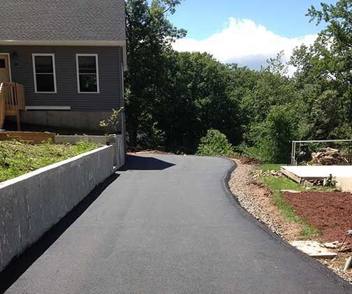 Asphalt driveway curves toward a gray house next to a concrete wall.