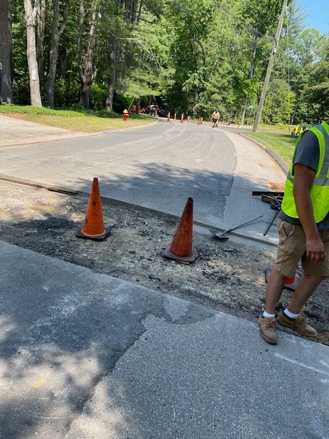 Road resurfacing: worker in vest near cones, section of street torn up, with fresh asphalt in the background.