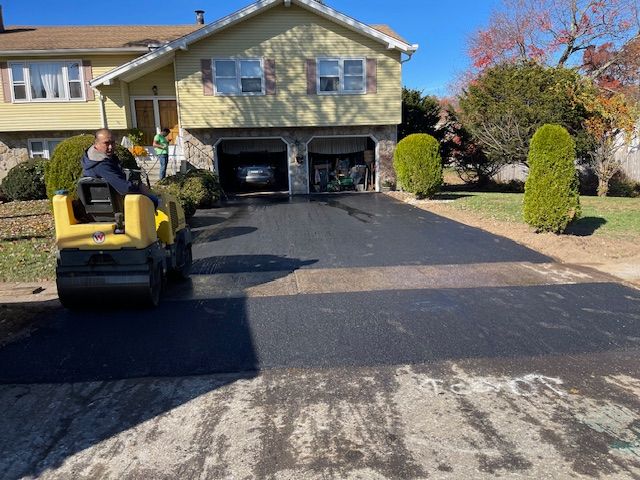 Man operating asphalt roller paving a driveway in front of a two-story house.