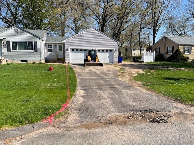 Driveway being worked on with asphalt damage, a small bobcat and a house in the background.