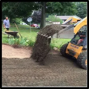 A skid steer dumping dirt onto a driveway. A man watches nearby. Outdoors, daylight.