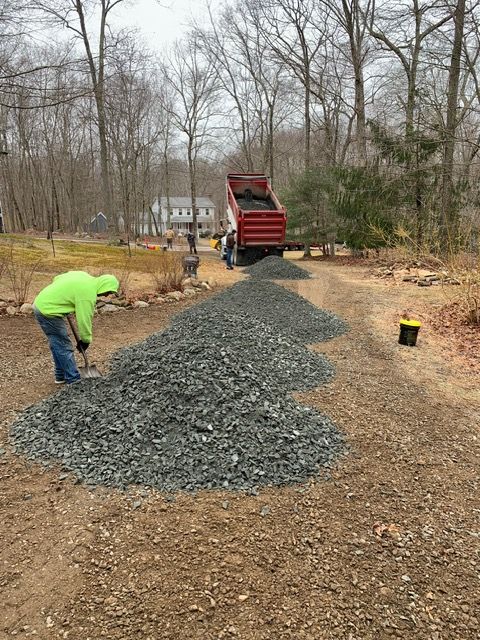 Person in neon green jacket spreading gravel from a dump truck on a driveway.