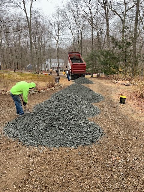 A worker in neon green jacket spreads gravel from a dump truck onto a driveway.