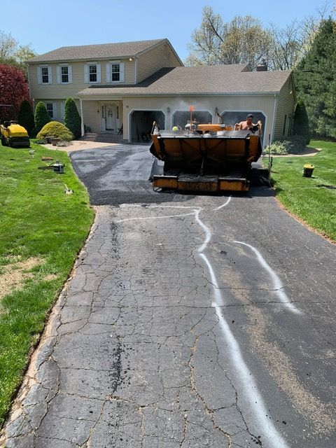 Driveway resurfacing: paving machine on cracked asphalt driveway in front of a two-story house.