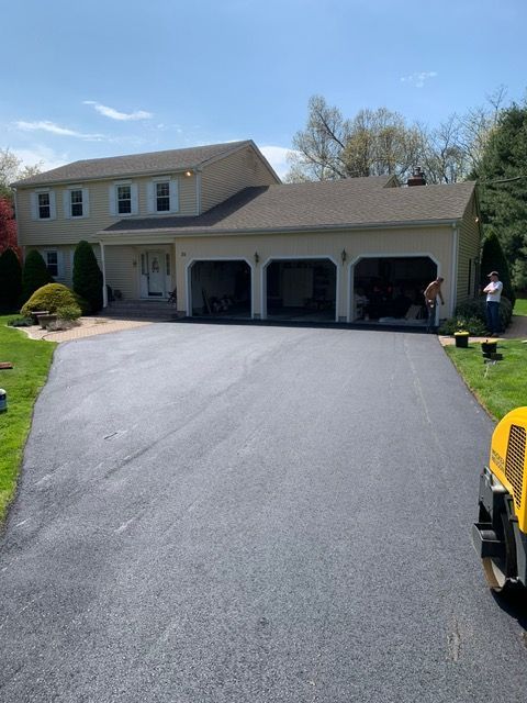 A newly paved asphalt driveway leads to a two-story house with a three-car garage. Two people are near the garage.