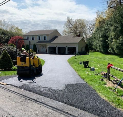 Asphalt paving driveway; person operating a roller machine; house with garage in the background.