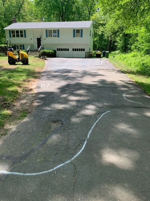 Asphalt driveway leading to a two-story white house, marked with white lines, construction equipment nearby.