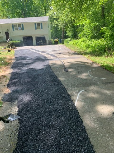 New black asphalt driveway being laid next to an older concrete driveway, in front of a two-story house.
