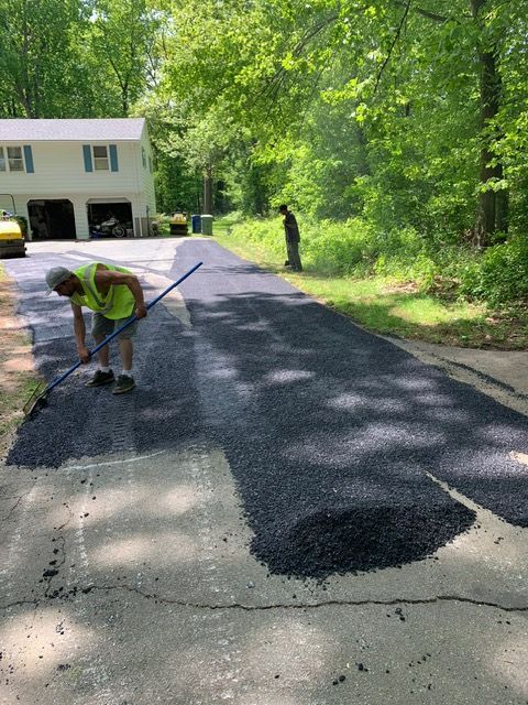 Man paving a driveway with asphalt. He’s using a long tool. Another person stands nearby.