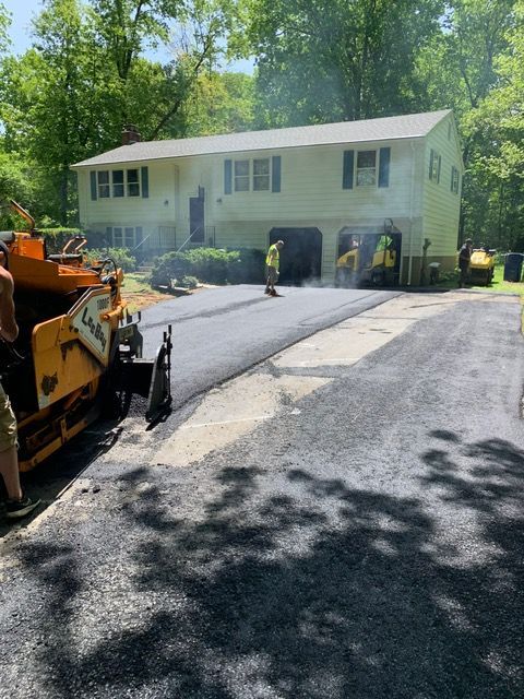 Workers paving a driveway in front of a light-colored two-story house on a sunny day.