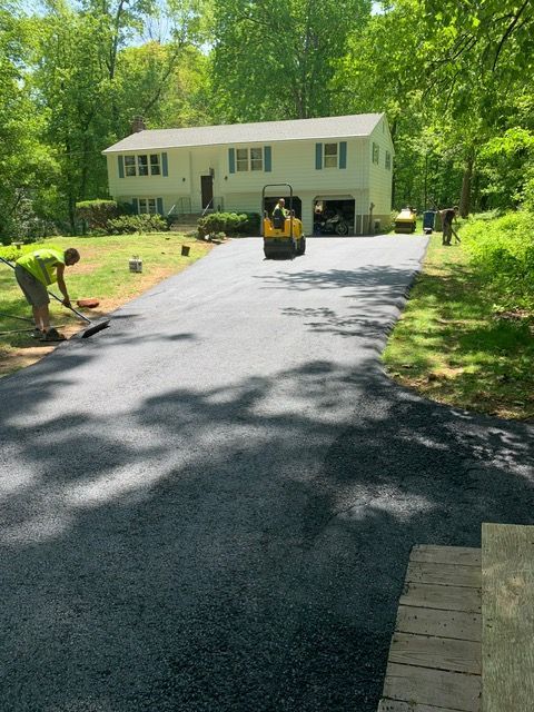 Asphalt driveway paving: workers with roller and rake, house in background.