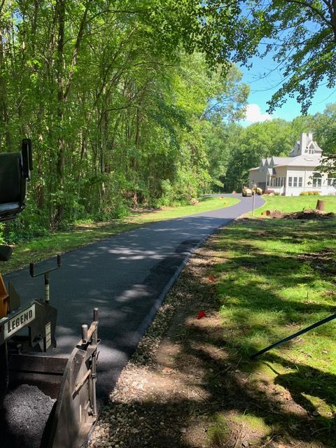 Freshly paved asphalt driveway leading to a house surrounded by trees on a sunny day.