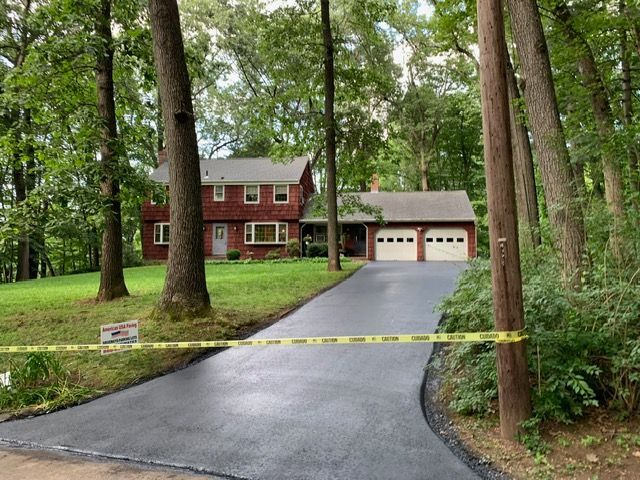 A two-story brick house with a freshly paved driveway, surrounded by trees. Yellow caution tape is across driveway.