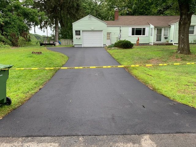 Newly paved black asphalt driveway leading to a light green house and garage, green grass on both sides.