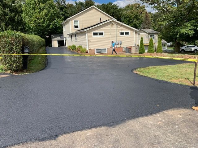Newly paved asphalt driveway curving towards a two-story beige house with a person walking.