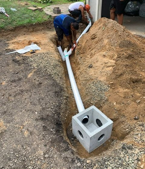 Construction workers installing a drainage system with concrete catch basin, pipes, and excavated trench.