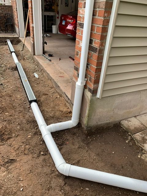 White drainage pipes diverting water away from a garage. Installation in progress, visible trench and dirt.