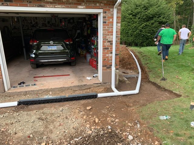 Workers installing a drainage system next to a garage. A car is inside the garage.