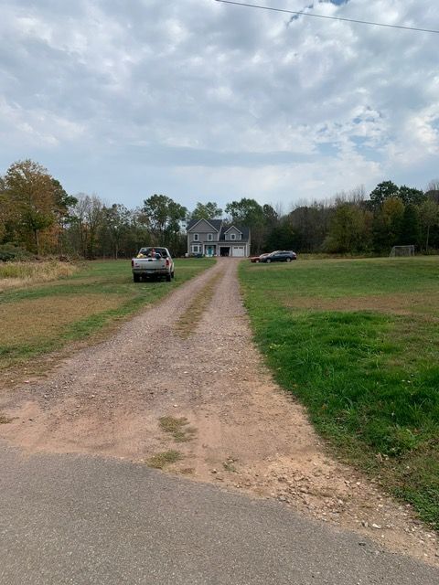 Gravel driveway leading to a gray house, cars parked on either side, set in a grassy yard under a cloudy sky.