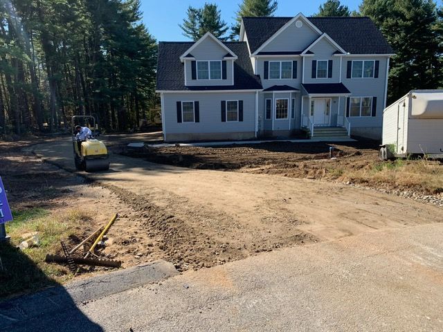 Driveway construction in front of a two-story house; a roller compacts the dirt.