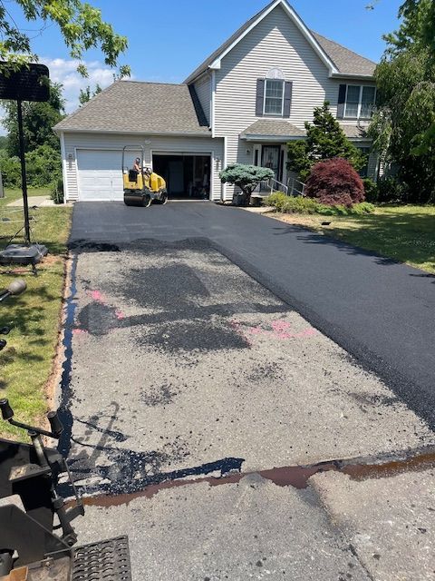 Asphalt driveway paving in progress; roller compacting new asphalt, home in the background.