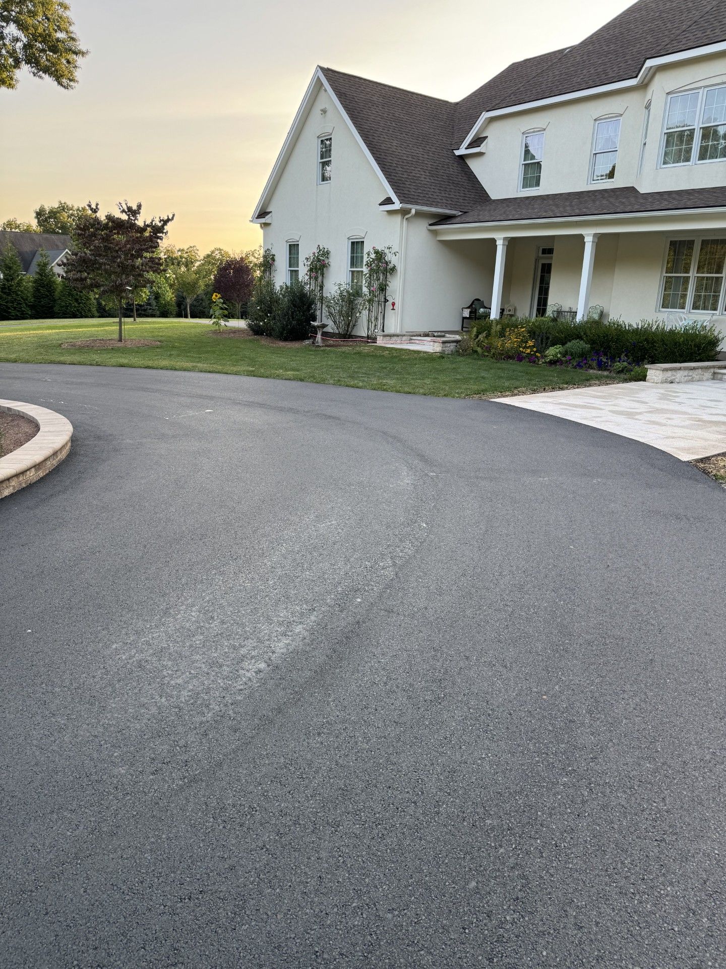 Black asphalt driveway curves toward a two-story beige house with a porch. Sunset.