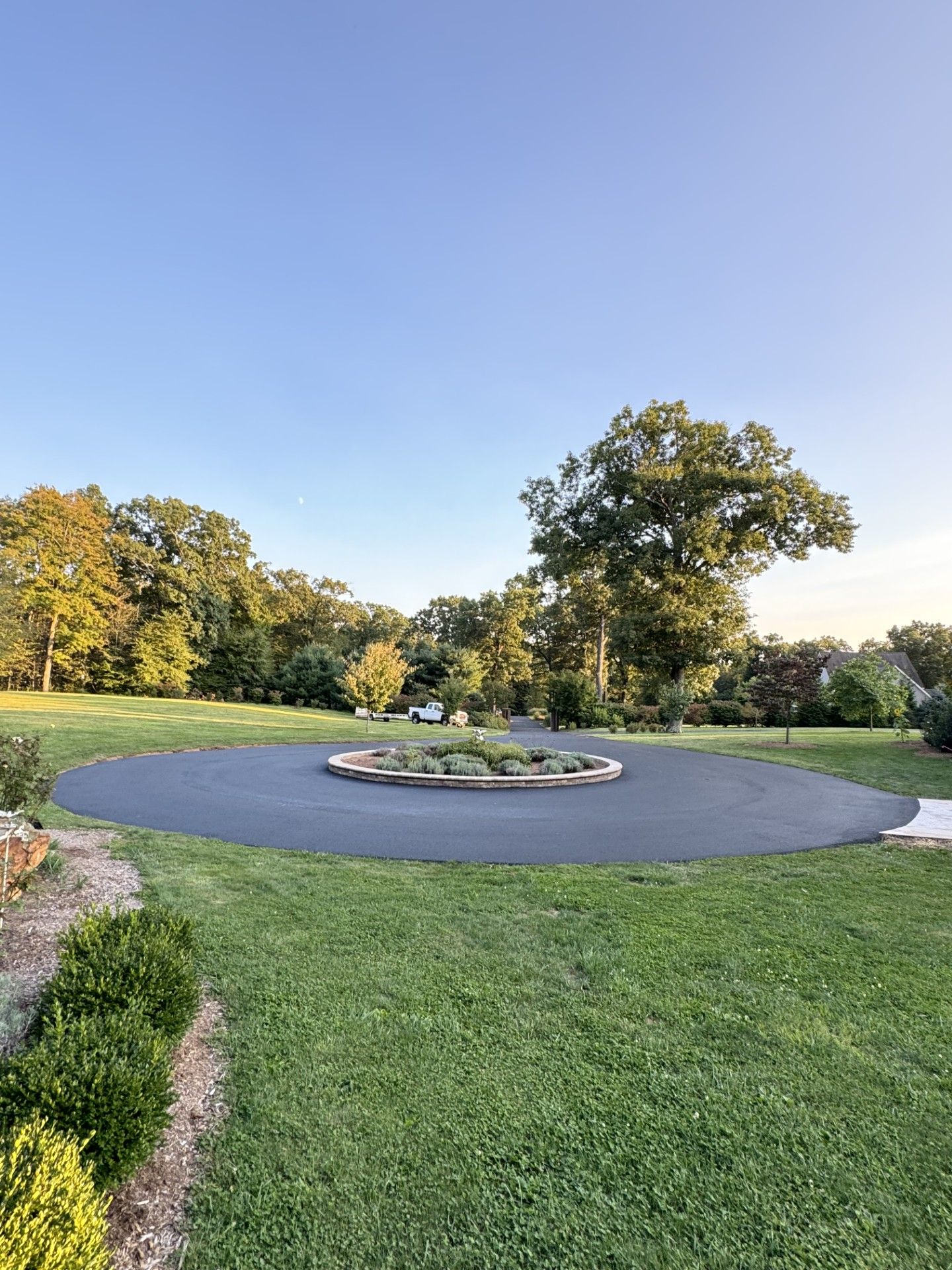 Circular asphalt driveway with central garden bed, surrounded by green lawn and trees under a blue sky.