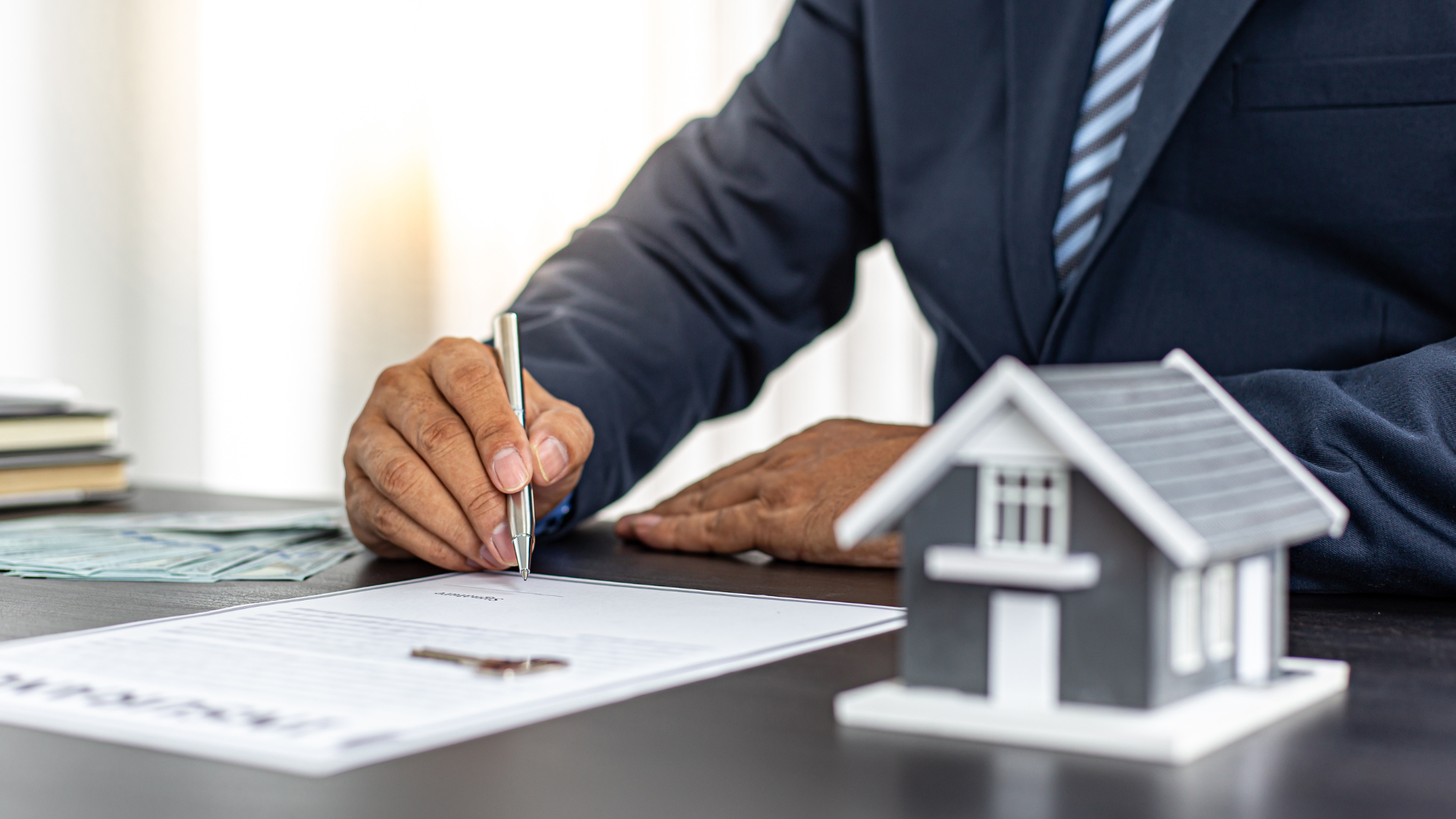 A businessman signs documents beside a small house model on a desk.
