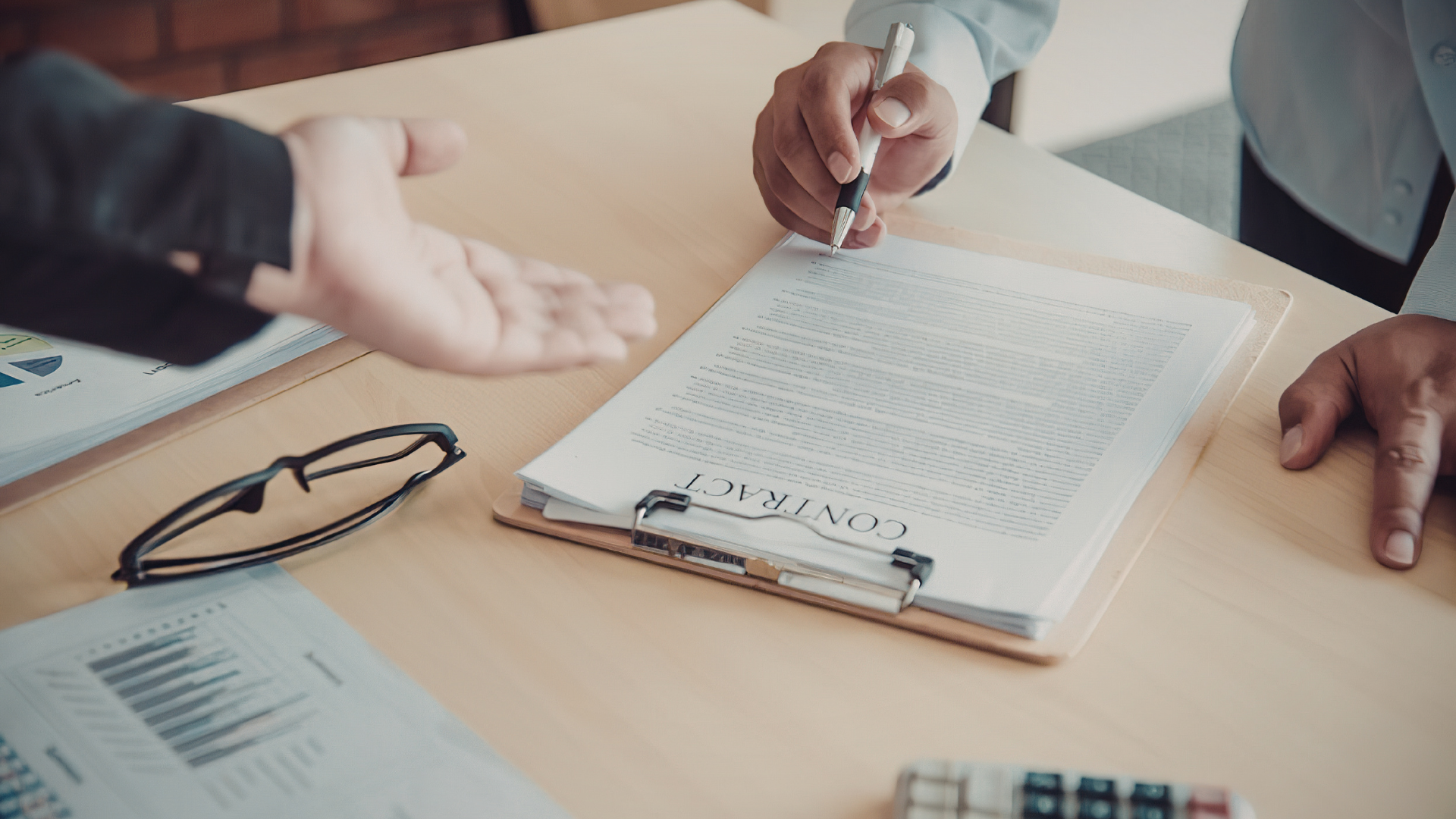 Hands reviewing paperwork on a conference table with glasses and a pen on a laptop nearby