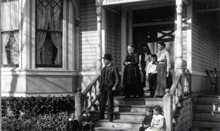 People standing on a porch steps in front of a house, with curtains in the windows and a child seated at the bottom steps