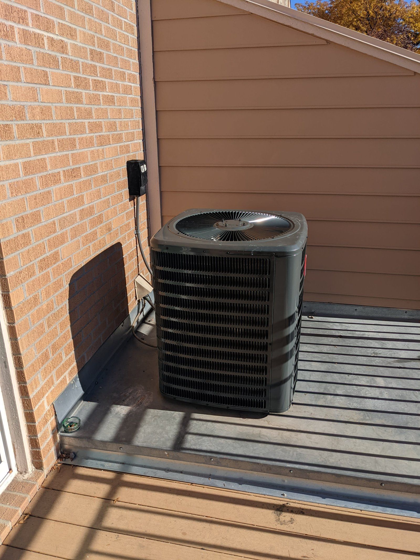 Air conditioning unit on a deck next to a brick wall and siding.