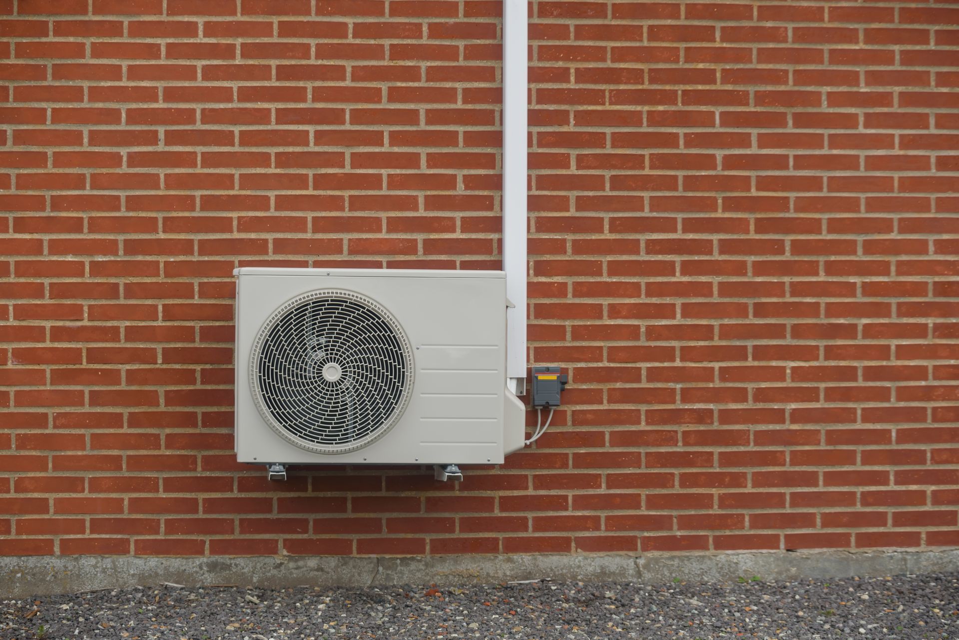 Air conditioning unit mounted on a red brick wall. White pipes run upwards.