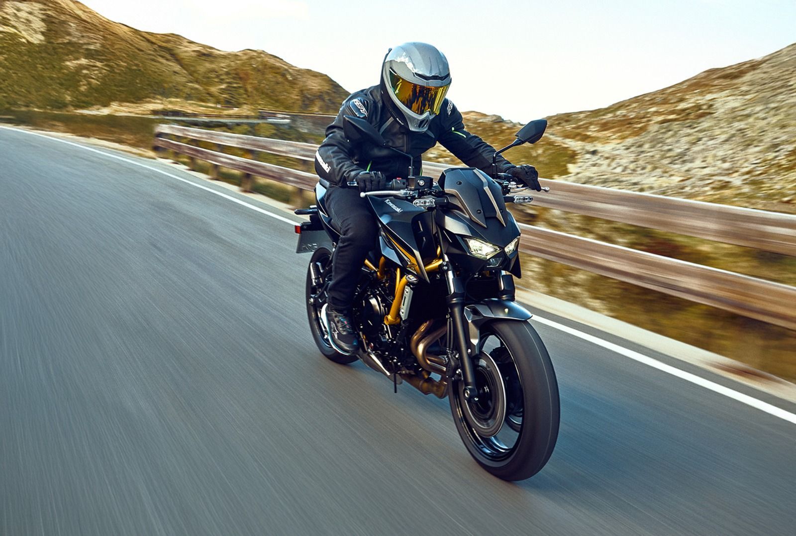 Motociclista de negro circulando por una carretera con curvas a través de un paisaje montañoso.