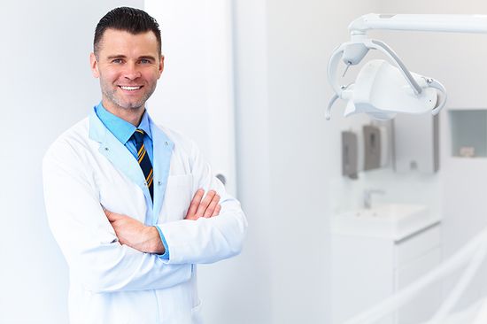A dentist in a white coat and blue shirt with a tie stands smiling with arms crossed in a bright, modern dental office.