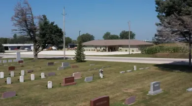 Small cemetery with scattered gravestones on a grassy lawn beside a road and low buildings under a blue sky