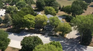 Aerial view of a tree-lined suburban yard with a house, driveway, and road nearby
