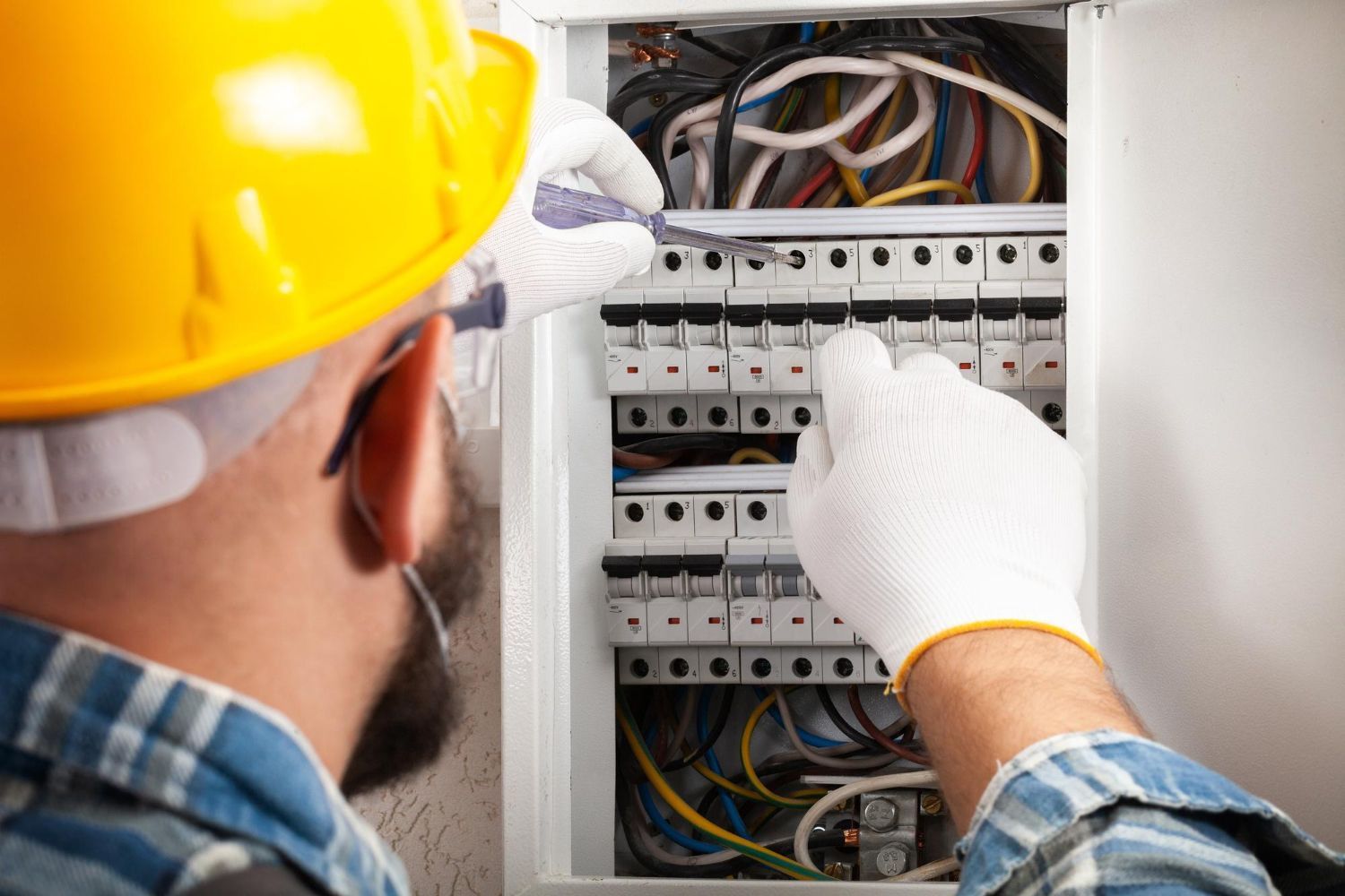 An electrician in a yellow hard hat and white gloves inspecting a circuit breaker panel.