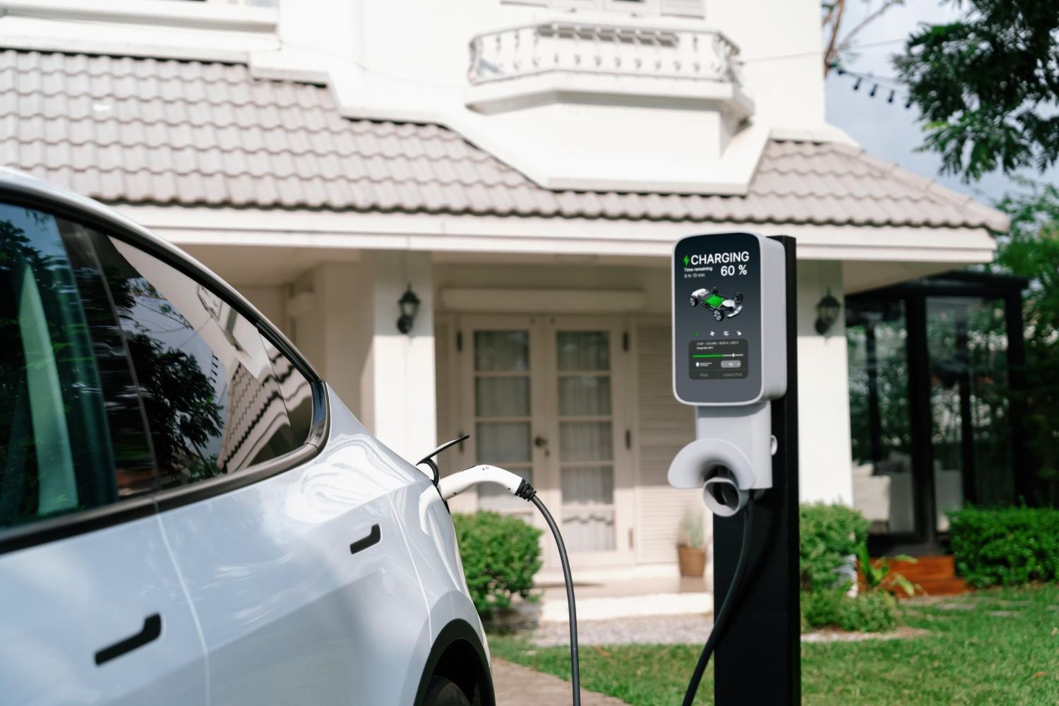 A white electric vehicle parked at a home, charging via an EV station with a digital screen on a black pedestal.