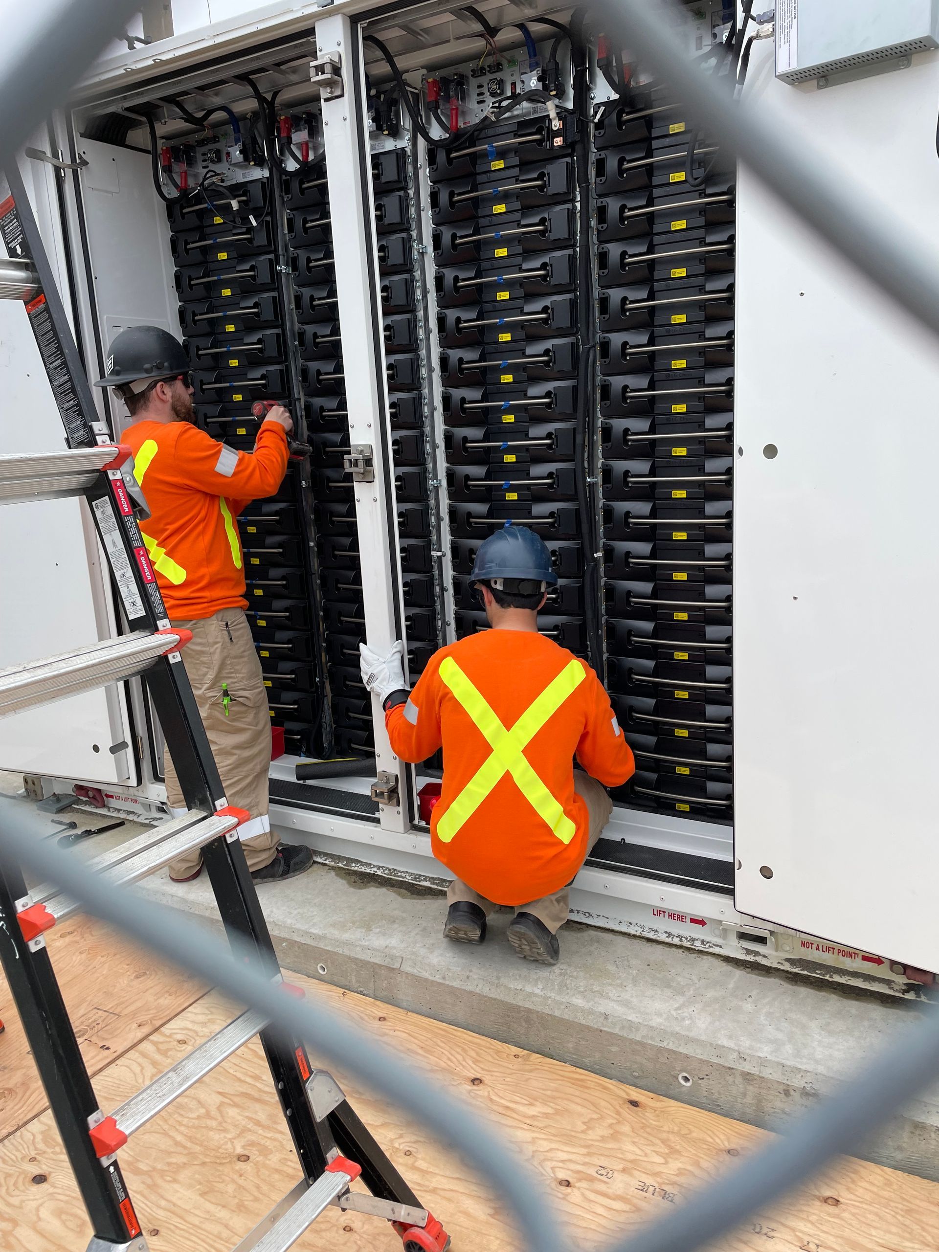 Two men are working on a large electrical box behind a fence.