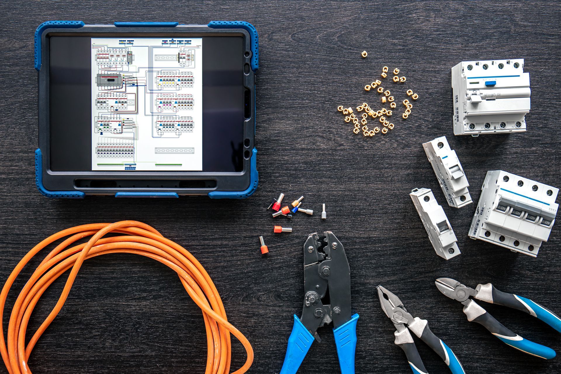 Tablet displaying electrical schematic, surrounded by electrical tools and components on dark surface.
