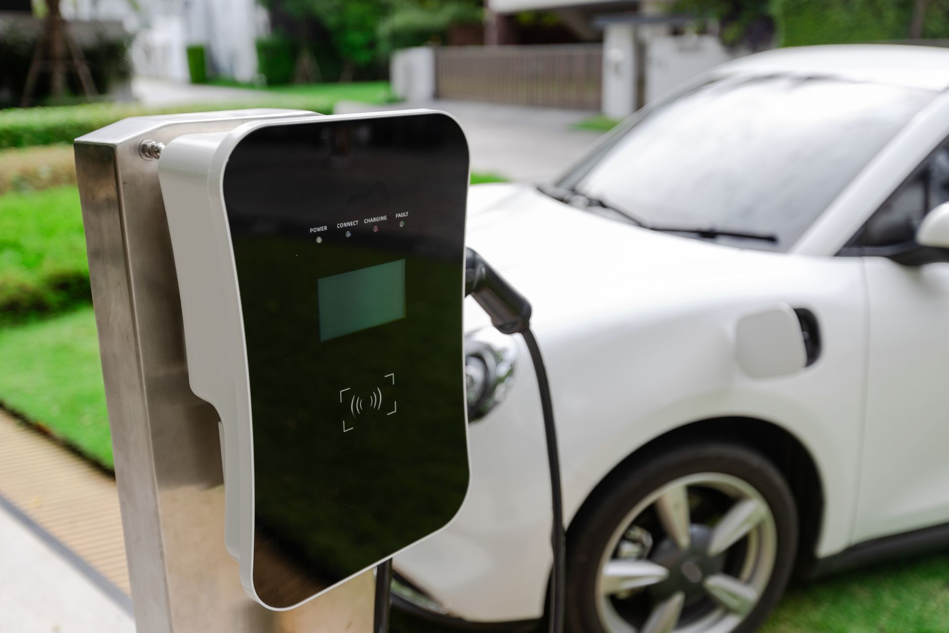 White electric car charging at a home charging station. Green grass and house in the background.