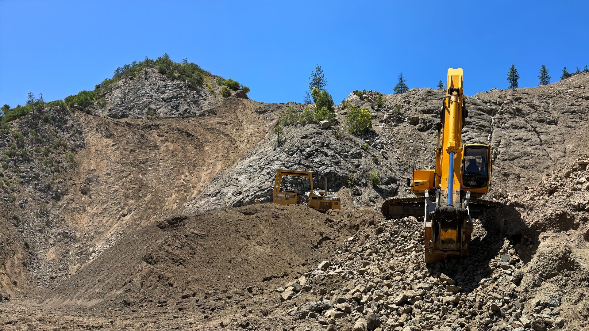 A large yellow excavator is working on a rocky hillside in Ashland, Oregon.