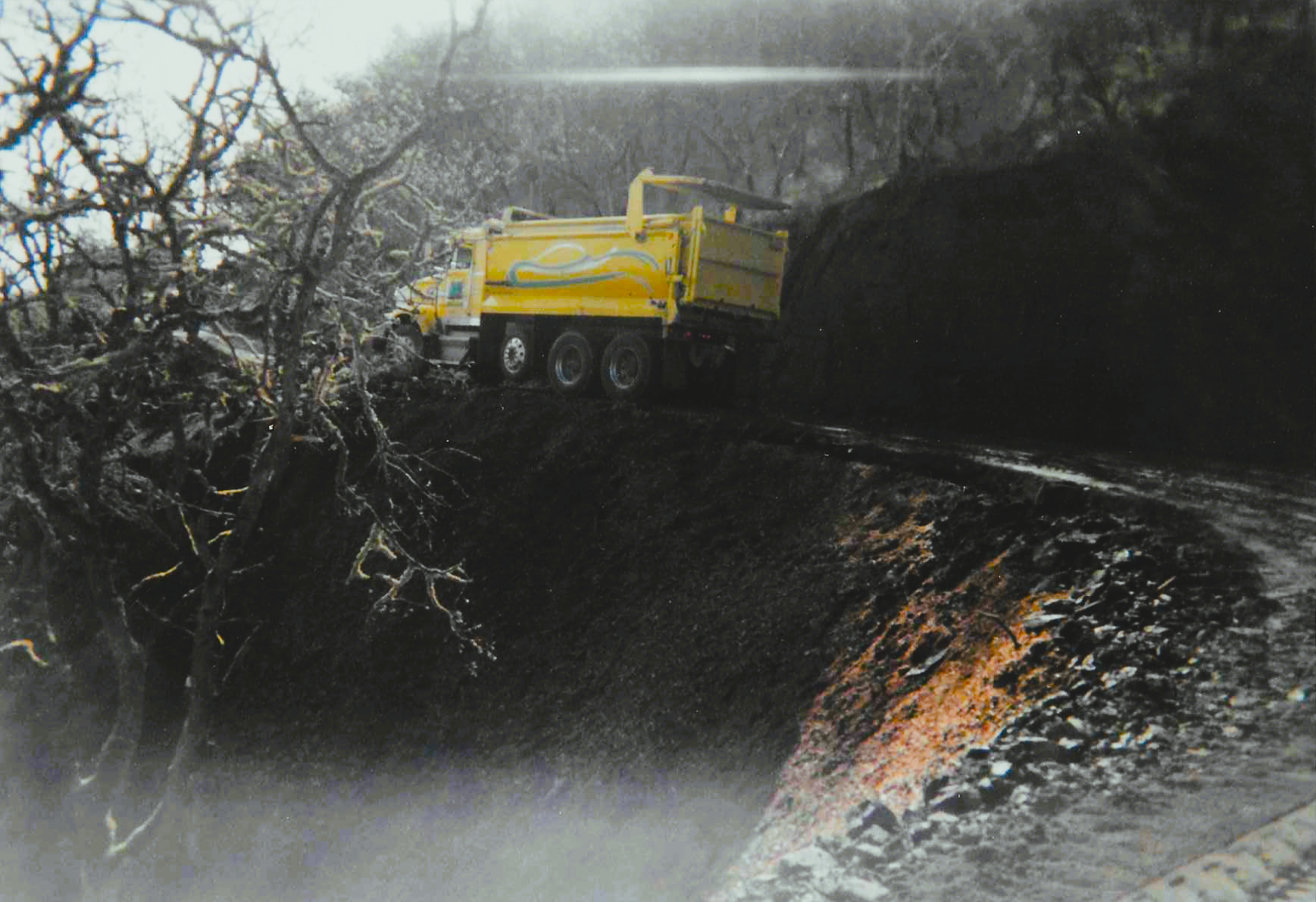 A yellow dump truck is driving down a dirt road in the woods of Ashland, Oregon.